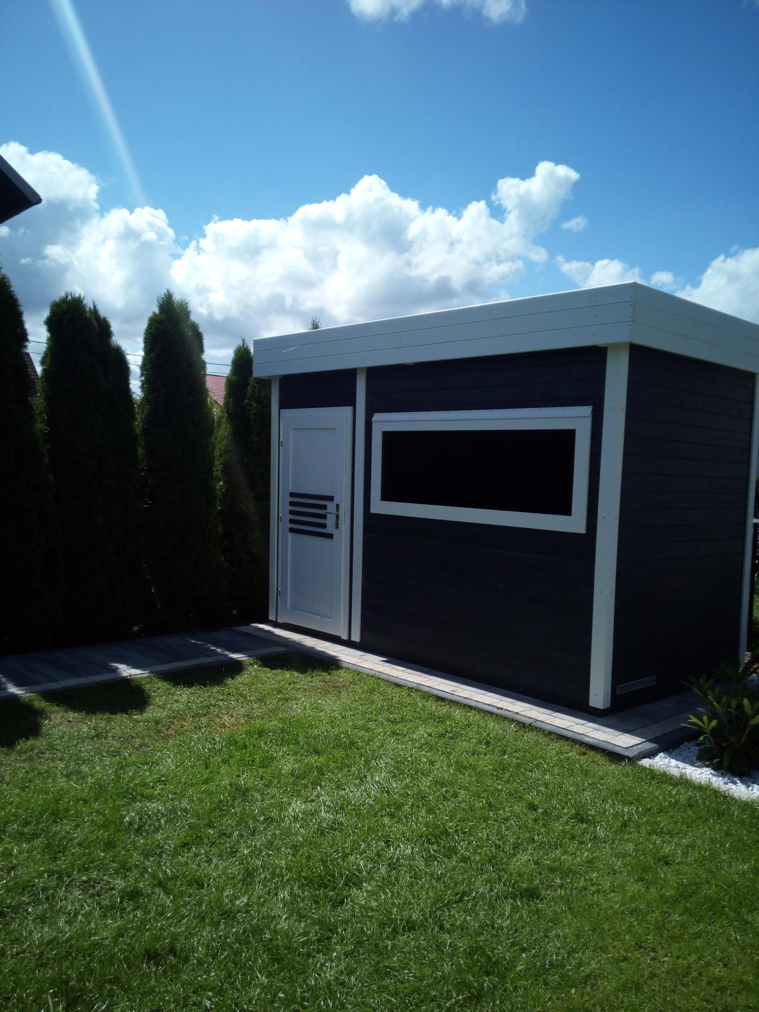 Nowoczesna, ciemnoszara altana ogrodowa z białymi detalami, widoczna z boku na tle zielonego trawnika, z tujami po lewej stronie i niebieskim niebem z chmurami w tle.