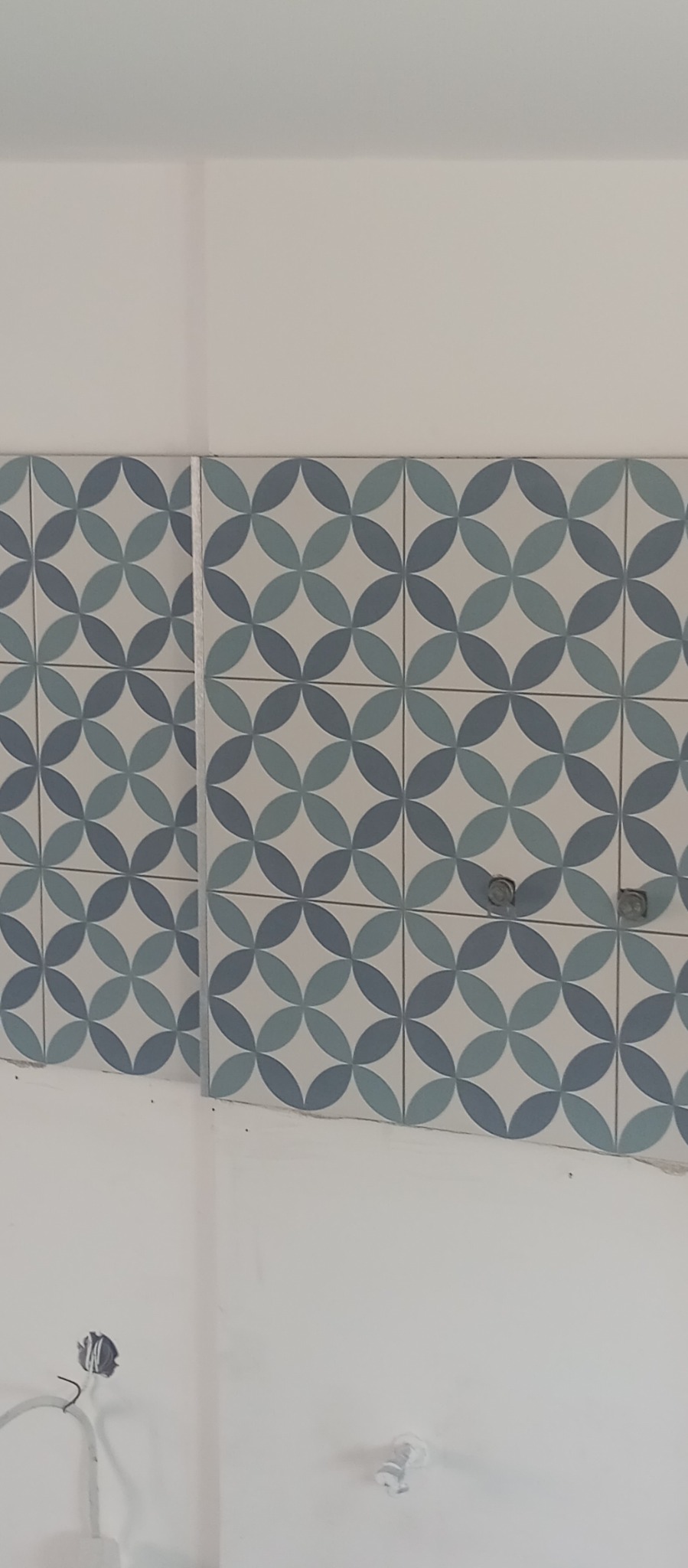 Ściana wyłożona dekoracyjnymi, błękitno-szarymi płytkami ceramicznymi z motywem kwiatowym. Widoczne gniazdka elektryczne i fragment instalacji.