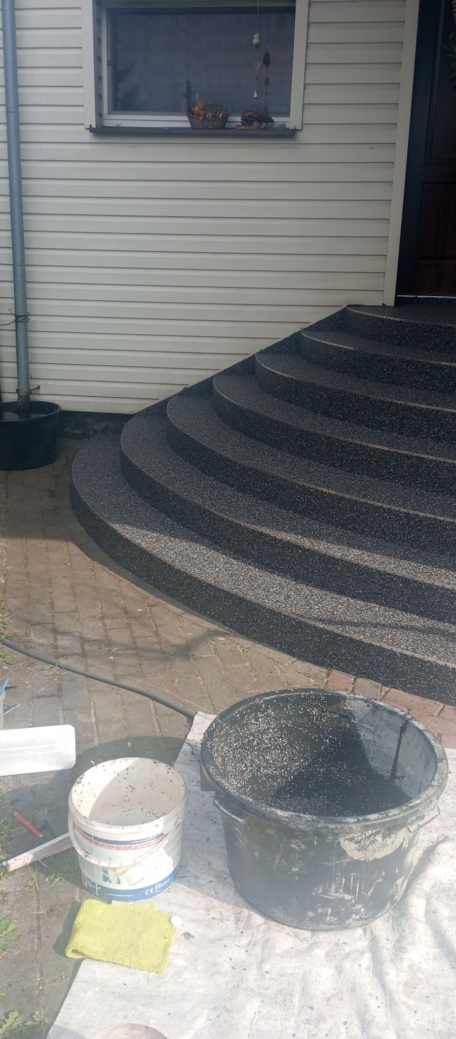 Wykończone schody wejściowe z żywicy epoksydowej w kolorze grafitowym, widoczne narzędzia i wiaderka po pracy. Elewacja budynku z sidingu.