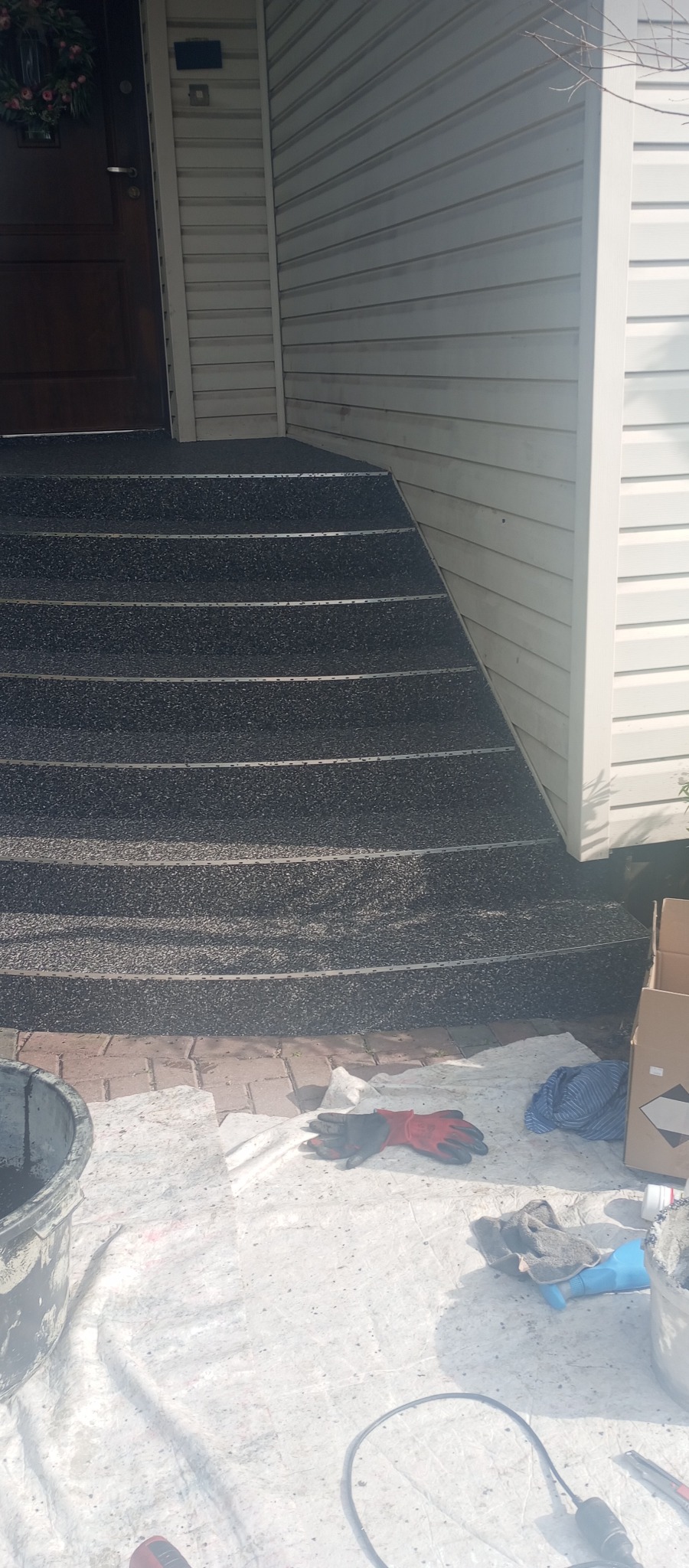 Wykończone schody wejściowe z szorstką, ciemną nawierzchnią i metalowymi listwami antypoślizgowymi, obok ściana z białym sidingiem i narzędzia pracy.