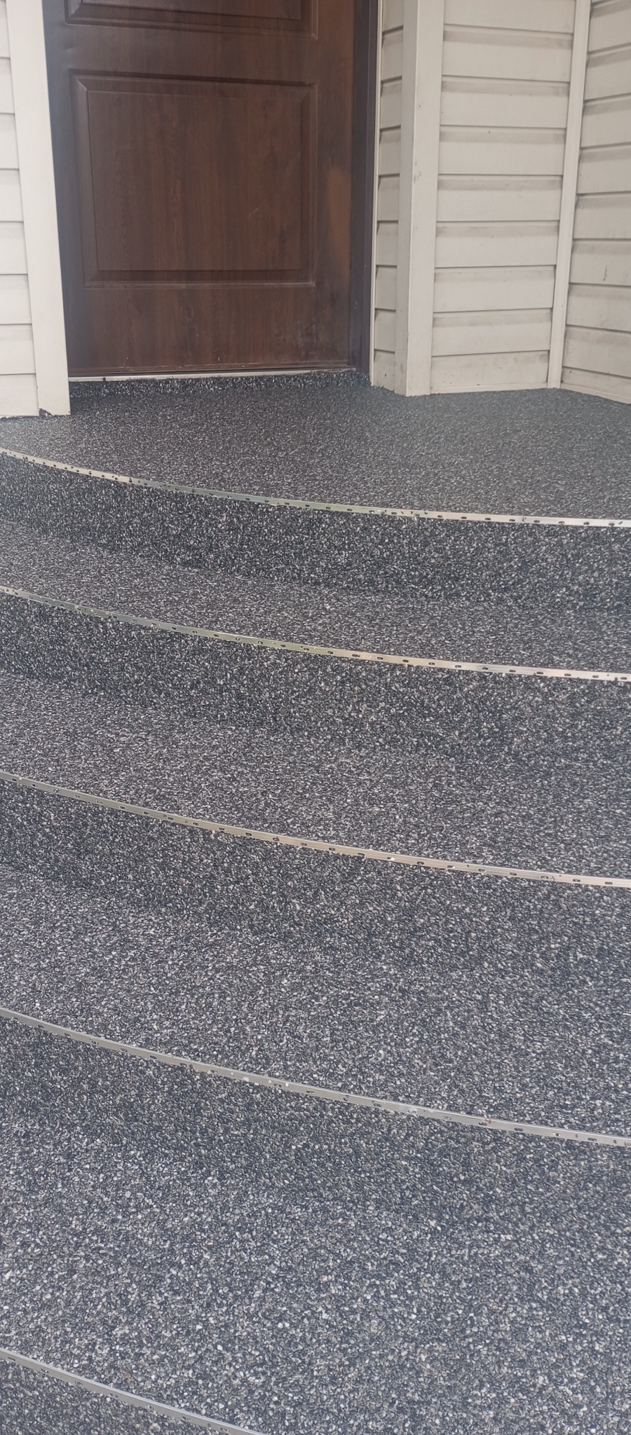 Wykończone schody wejściowe z szarą, kamienną nawierzchnią i metalowymi listwami, prowadzące do brązowych drzwi wejściowych z białym obramowaniem.