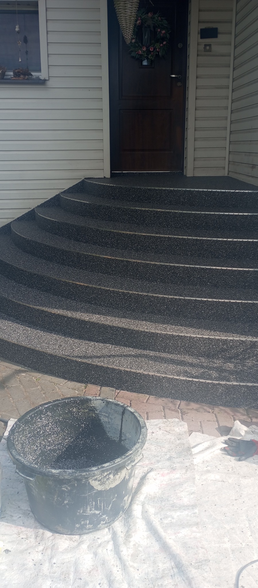 Wykończone schody wejściowe do domu z żywicy epoksydowej w kolorze grafitowym. Widoczne detale wykończenia i wiadro z resztkami materiału.