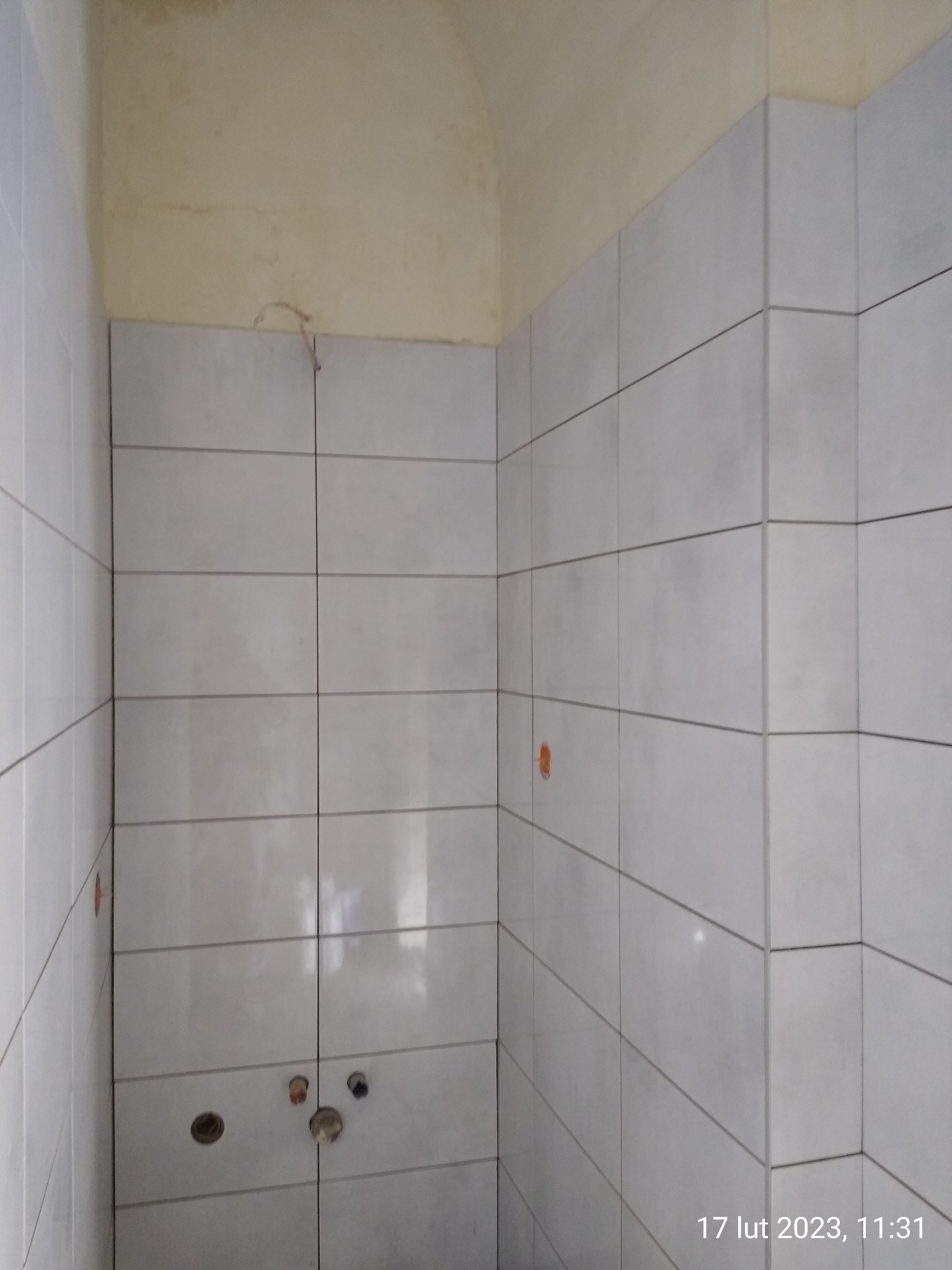 Ściany w łazience wyłożone jasnymi, prostokątnymi płytkami. Widoczne otwory na armaturę i niedokończone górne partie ścian. Minimalistyczny styl wykończenia.