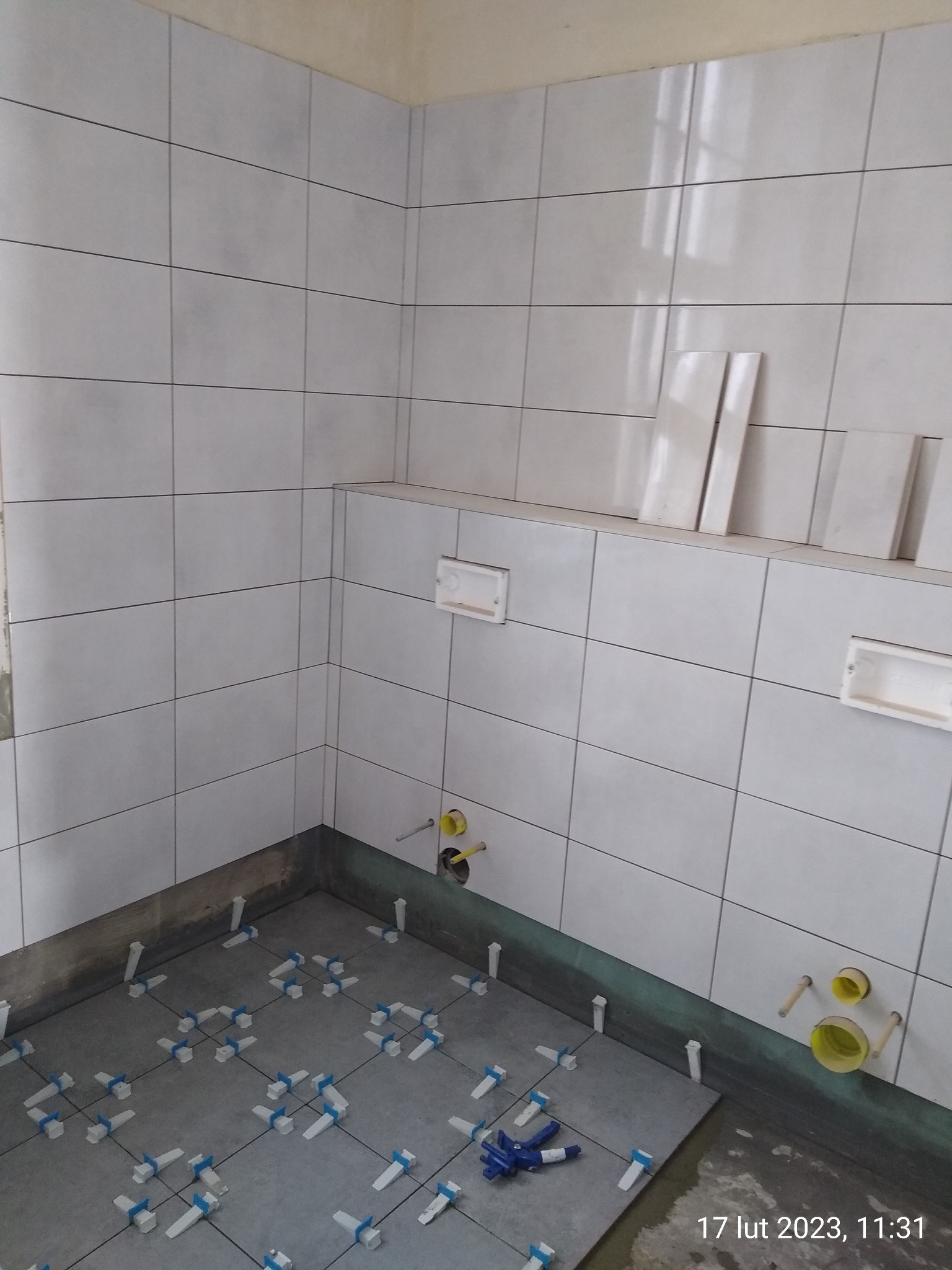 Układanie jasnych, kwadratowych płytek na ścianach i podłodze w łazience. Widoczne krzyżyki dystansowe i fragmenty nieułożonych płytek. Przygotowane podejścia wodne.