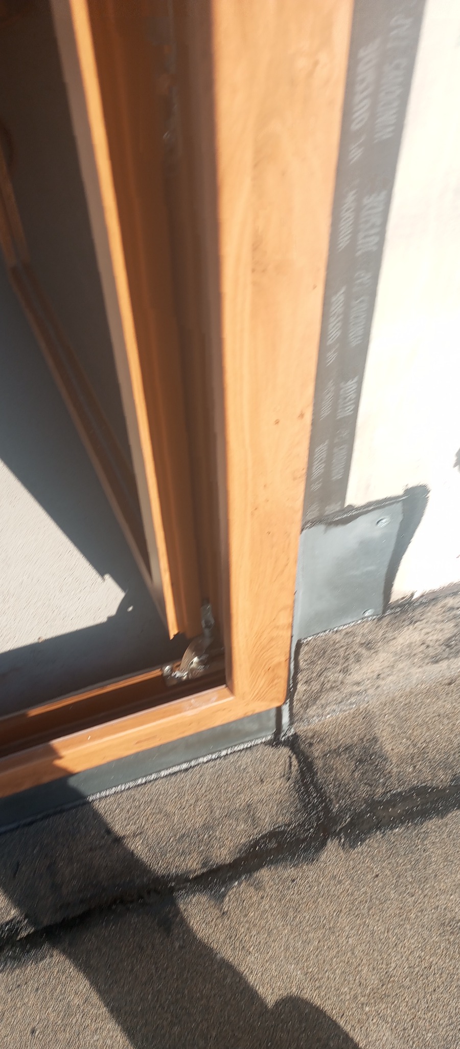 Fragment okna z drewnianą ramą, uszczelnionego taśmą izolacyjną i obróbką blacharską, zamontowanego w ścianie. Widoczne detale montażu i materiały.