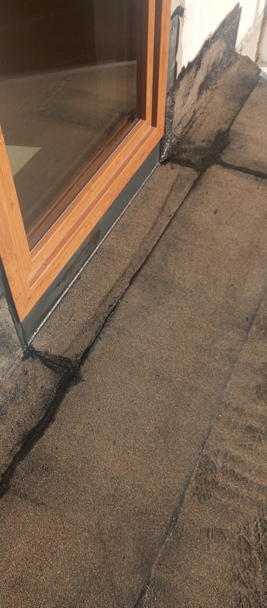 Szczegółowe ujęcie brązowej papy dachowej, przylegającej do drewnianej ramy okiennej. Widoczne łączenia papy i fragment ściany.