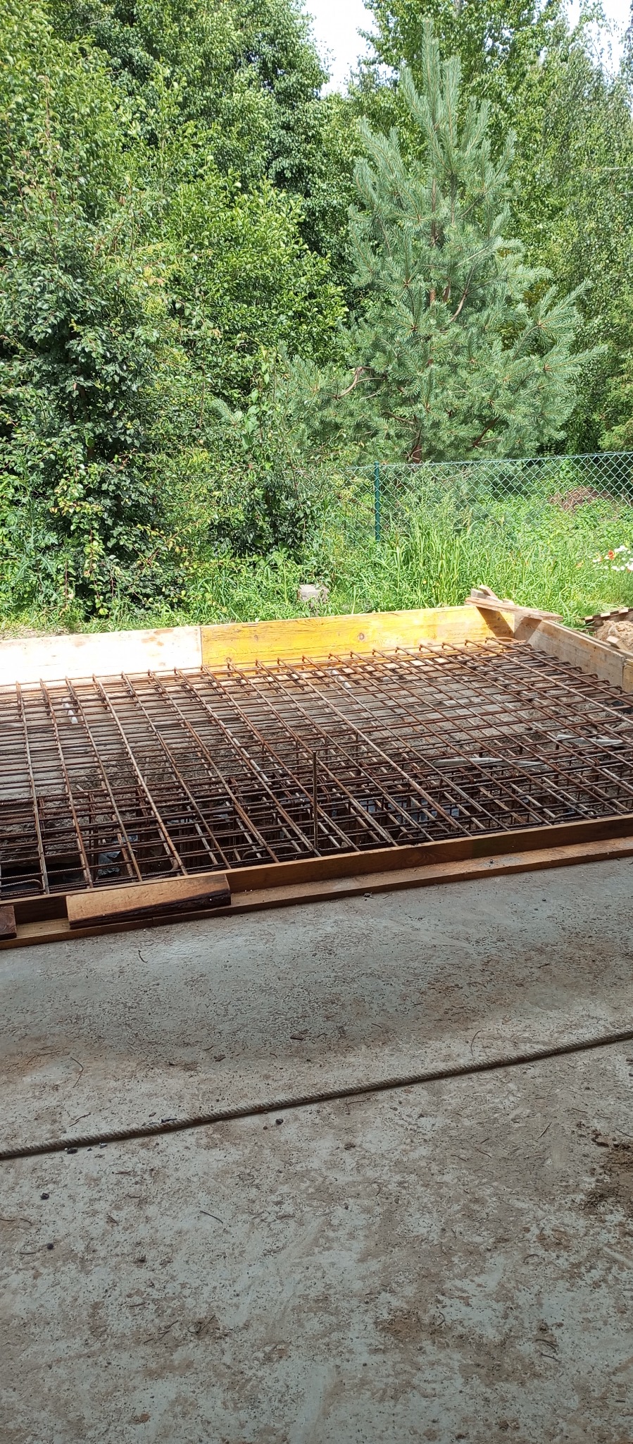 Zbrojenie fundamentów domu jednorodzinnego, widok z góry na siatkę z prętów stalowych w drewnianym szalunku, na tle zieleni drzew i krzewów.