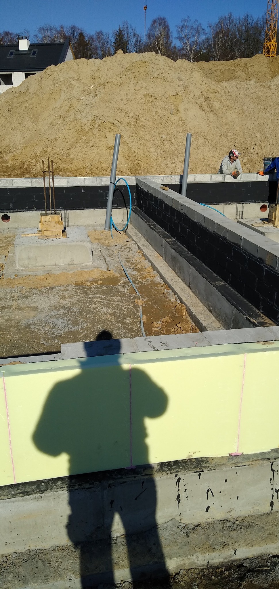 Fundamenty domu w trakcie budowy: izolacja pionowa, styropian, wystające rury. W tle hałda ziemi i częściowo widoczny budynek w stanie surowym otwartym.