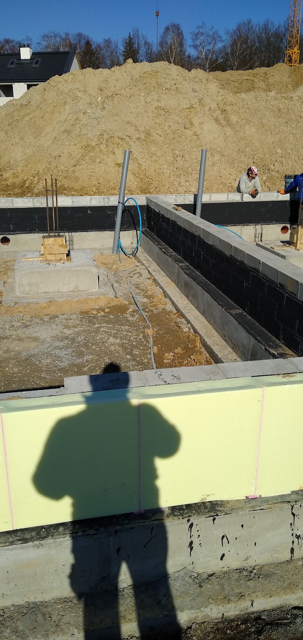 Fundamenty domu w trakcie budowy: izolacja termiczna styropianem, hydroizolacja, zbrojenie. W tle hałda ziemi i pracownik budowlany.