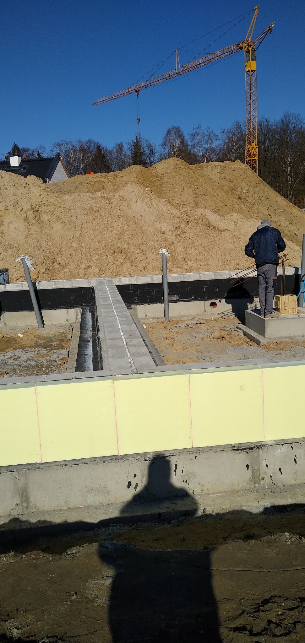 Fundamenty domu w trakcie budowy z widocznym ociepleniem, izolacją przeciwwilgociową i pracującym robotnikiem. W tle hałda piachu i żuraw budowlany.