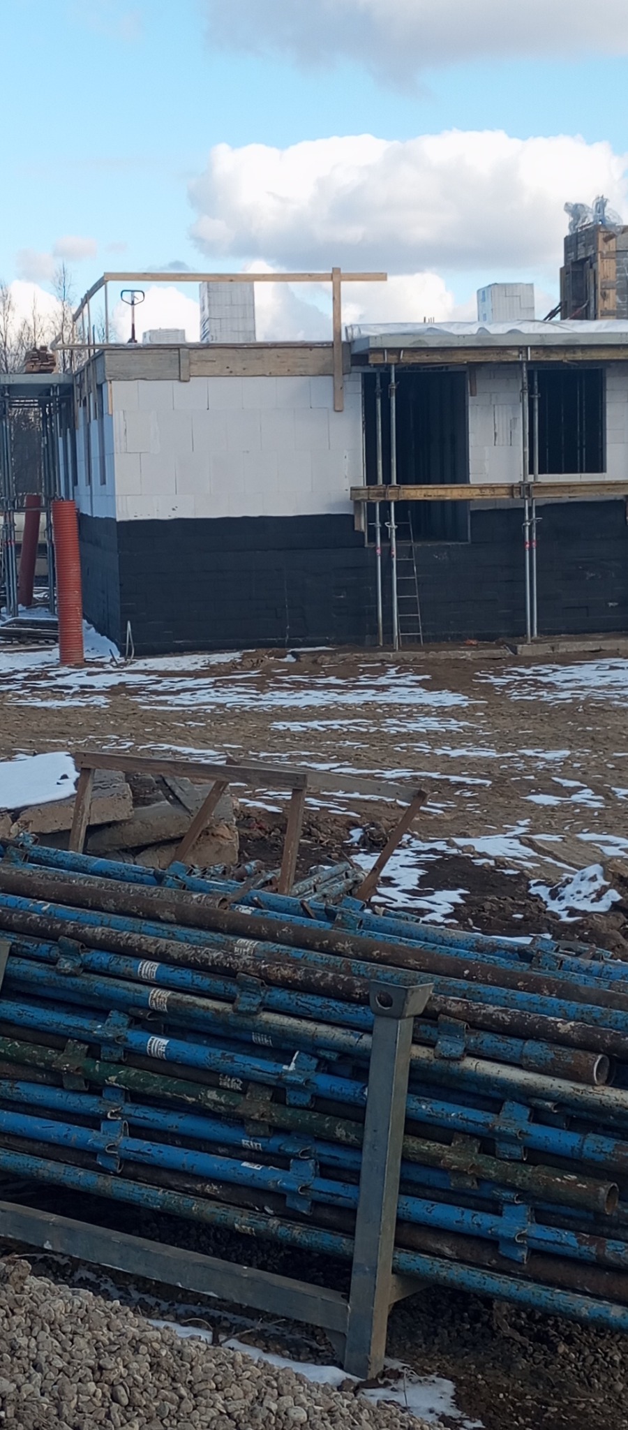 Budowa domu w trakcie: ściany z białego bloczka, czarna izolacja fundamentów, podpory, rusztowania. Na pierwszym planie stos niebieskich podpór budowlanych.