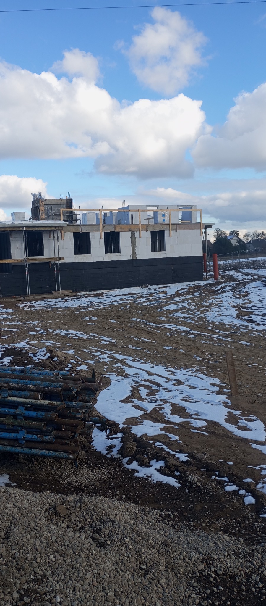 Budowa domu w stanie surowym otwartym, widoczne bloczki, czarna izolacja fundamentów, drewniane szalunki okienne, podpory budowlane i resztki śniegu na placu budowy.