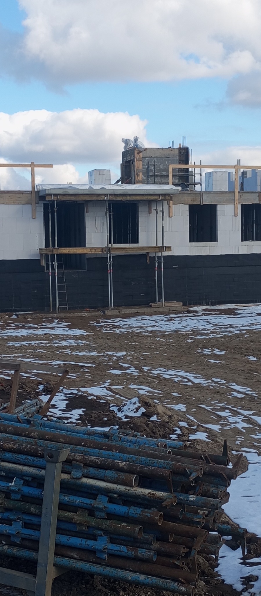 Budowa domu w trakcie realizacji: ściany z białych bloczków, czarna izolacja, rusztowania, podpory. Na pierwszym planie stos niebieskich podpór budowlanych.