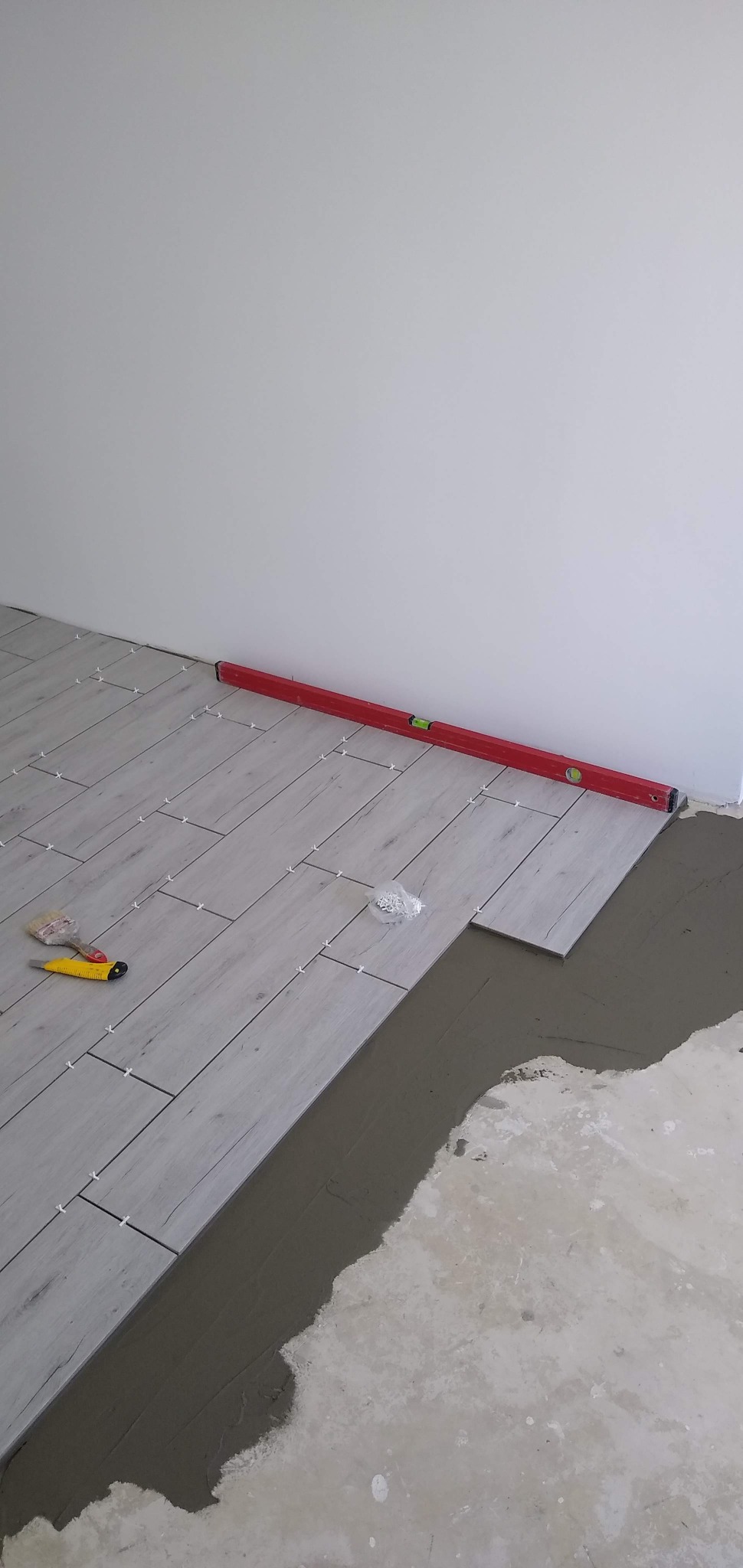 Układanie podłogi z płytek imitujących drewno. Widoczny poziomica, nóż do cięcia i krzyżyki dystansowe. Częściowo położone płytki na świeżym kleju.