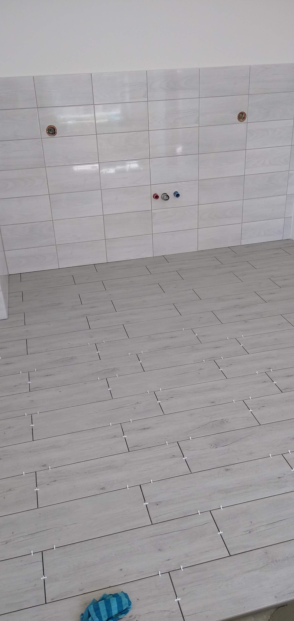 Świeżo położone, jasne płytki imitujące drewno na podłodze i ścianie w kuchni. Widoczne krzyżyki dystansowe i otwory na instalacje.