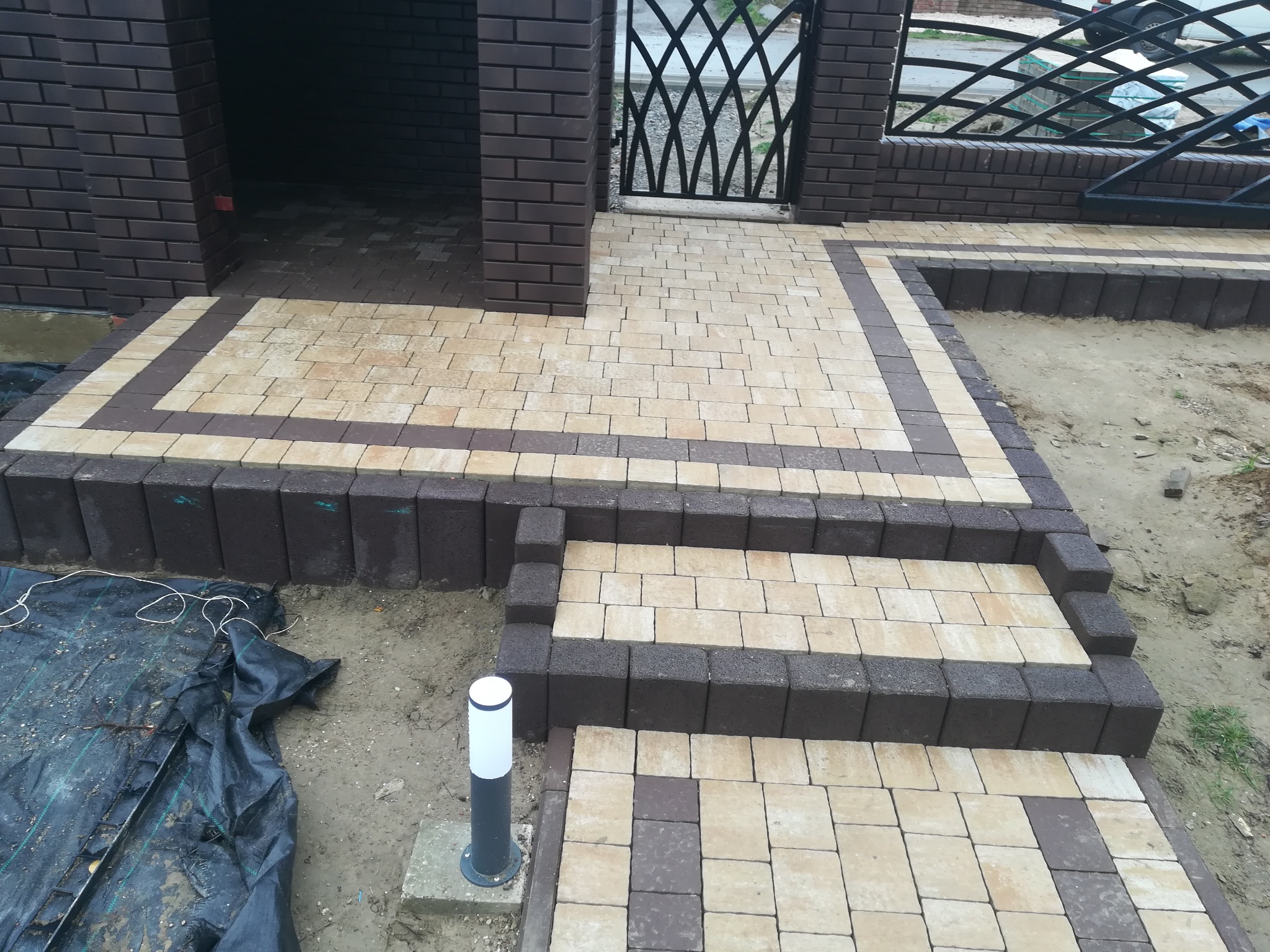 Ułożona kostka brukowa w dwóch odcieniach z obramowaniem, schodami i lampą ogrodową, widok na bramę i ceglany budynek