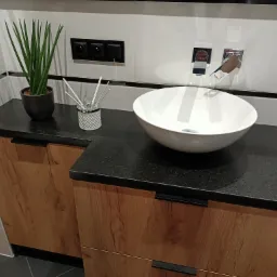 Czarny, nakrapiany blat z drewnianymi szafkami pod umywalką nablatową w nowoczesnej łazience, z dodatkami w postaci rośliny w doniczce i dyfuzora zapachowego.