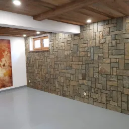 Wnętrze pomieszczenia z dekoracyjną ścianą wyłożoną kamiennymi płytkami o nieregularnych kształtach i odcieniach brązu i szarości, drewnianym sufitem z belkami i wpuszczonymi lampami, szarą podłogą...