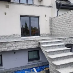 Zewnętrzne schody i taras wykończone szarą kamienną okładziną, widoczne detale tekstury kamienia i połączenia między elementami, w tle budynek z oknami.