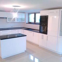 MeByl meble Bylica - Nowoczesna biała kuchnia z czarnym blatem, wyspa kuchenna z czarnym blatem, błyszczące fronty szafek, okno z drewnianą ramą, płytki podłogowe z połyskiem.