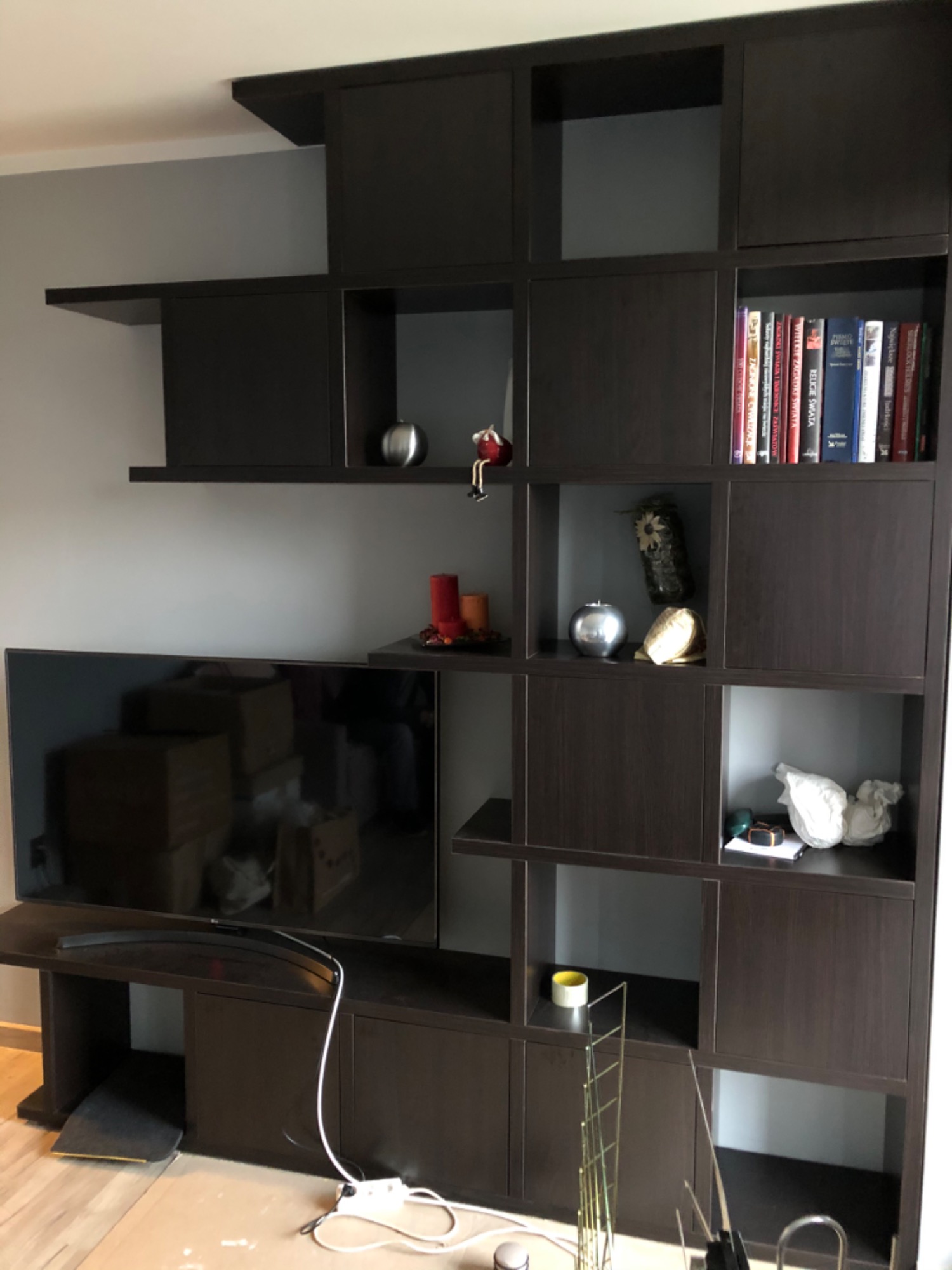 Ciemnobrązowy regał z otwartymi półkami, na których znajdują się książki, dekoracje i telewizor. Widoczne kable i przedłużacz na podłodze.