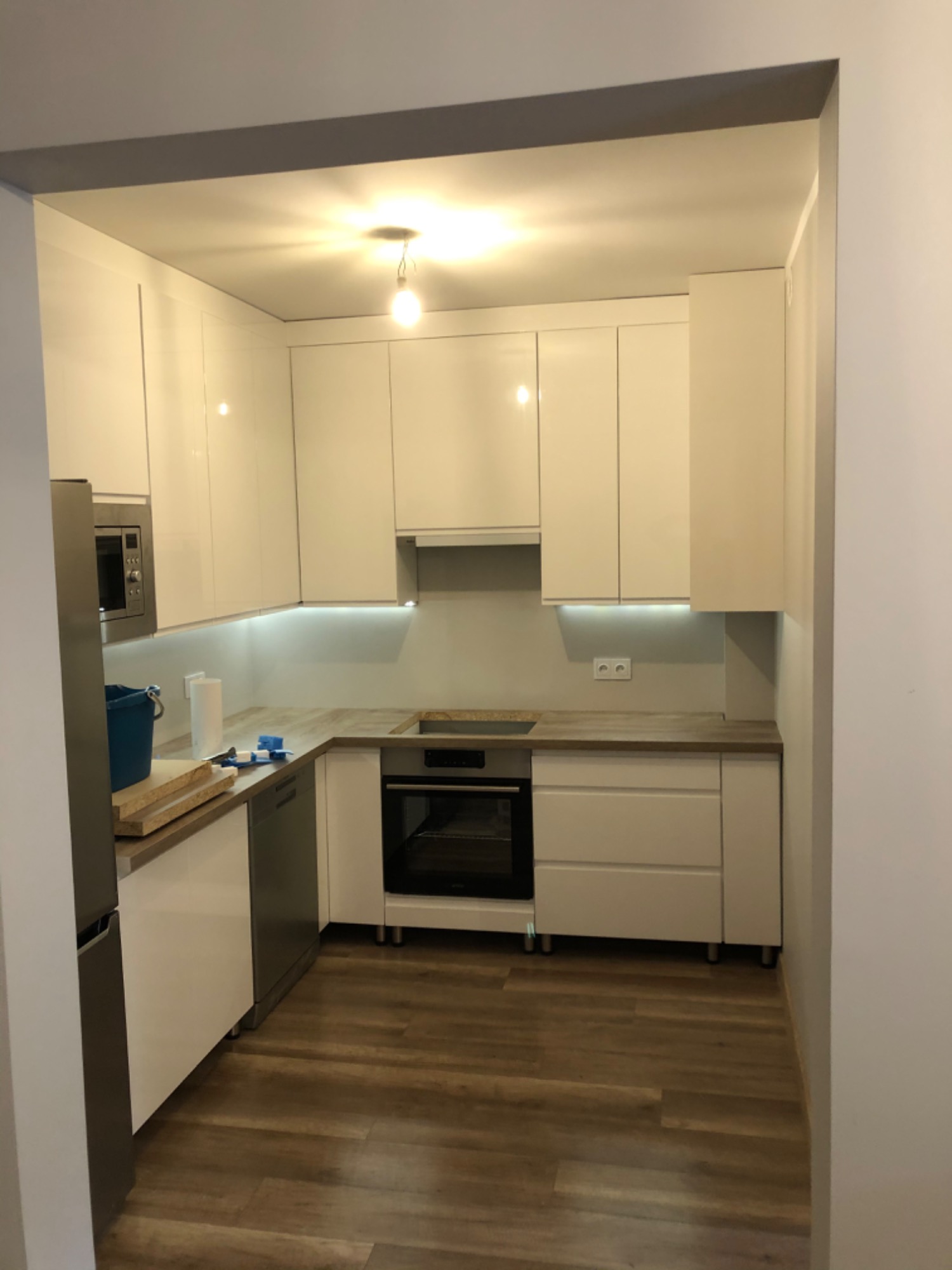 Nowoczesna, biała kuchnia z połyskiem, oświetleniem LED podszafkowym i drewnopodobnymi blatami, widziana z perspektywy wejścia do pomieszczenia, z widocznymi akcesoriami i narzędziami na blacie.