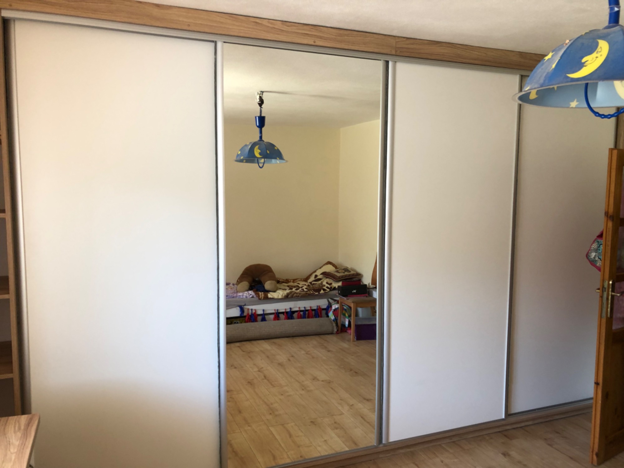 Biała szafa przesuwna z lustrem w pokoju dziecięcym, widoczne odbicie łóżka i lampki nocnej.