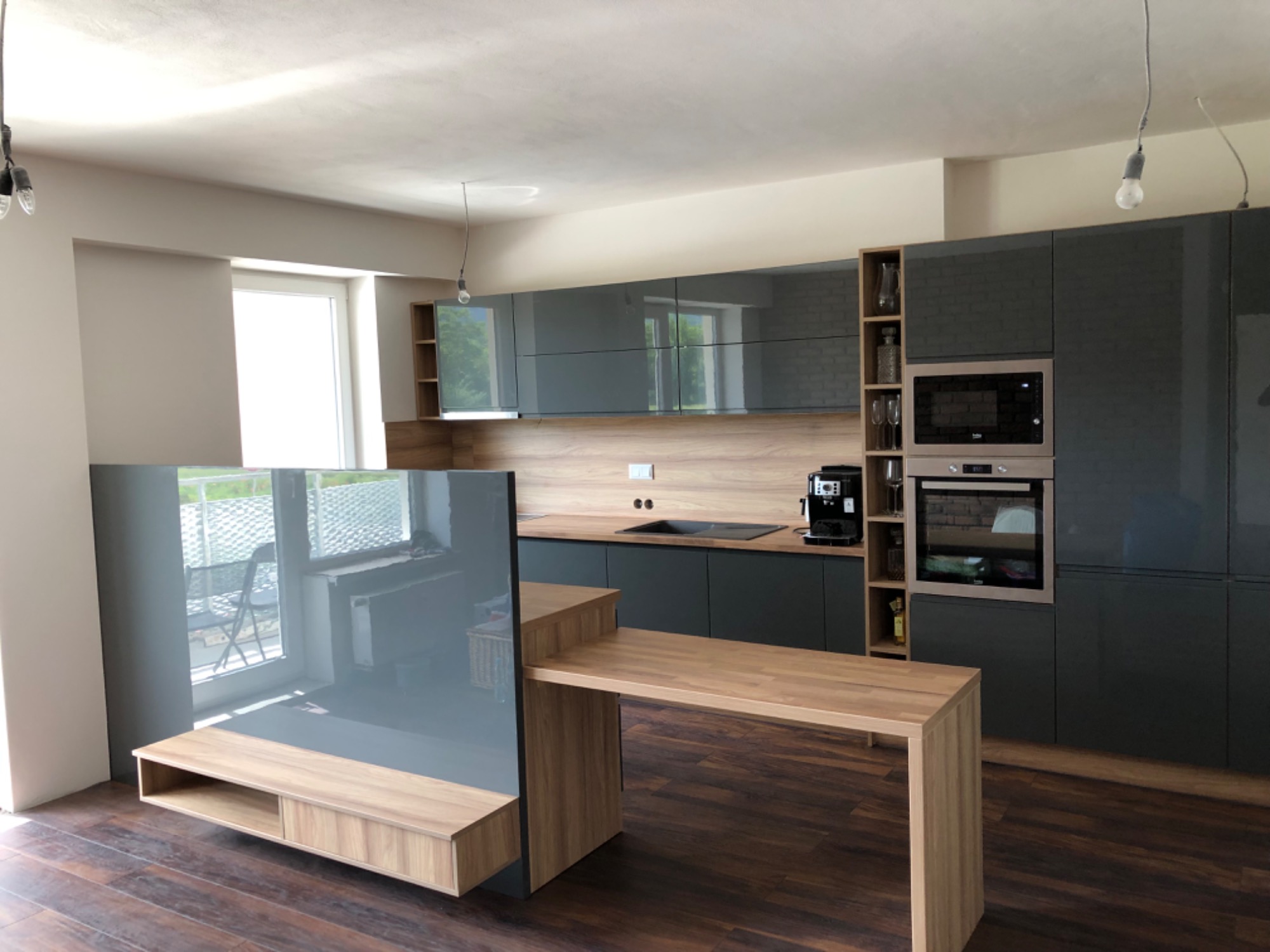 Nowoczesna kuchnia z szarymi, lakierowanymi frontami, drewnianym blatem i barkiem, z zabudowanym piekarnikiem i kuchenką mikrofalową, oświetlona wiszącymi żarówkami bez kloszy.