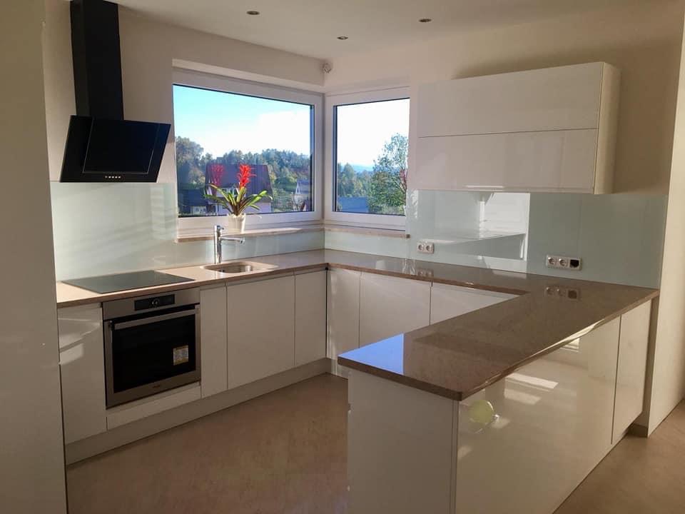 Nowoczesna, biała kuchnia na wymiar z blatem w odcieniach beżu, widok na zieleń za oknem, minimalistyczny design.