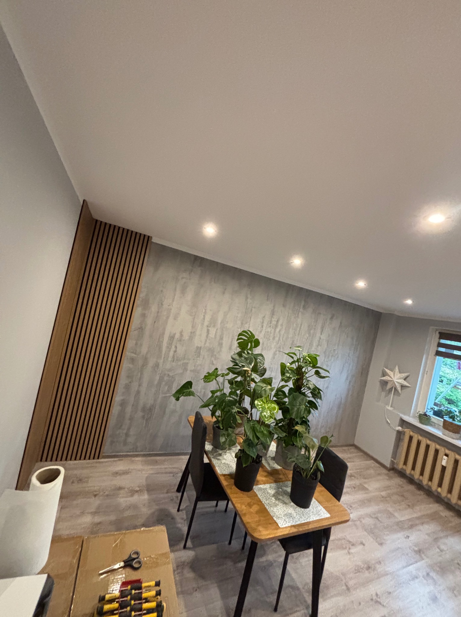 Nowoczesne wnętrze z panelami ściennymi z drewna, szarą ścianą imitującą beton i roślinami doniczkowymi na stole. Widoczne narzędzia sugerują prace wykończeniowe.