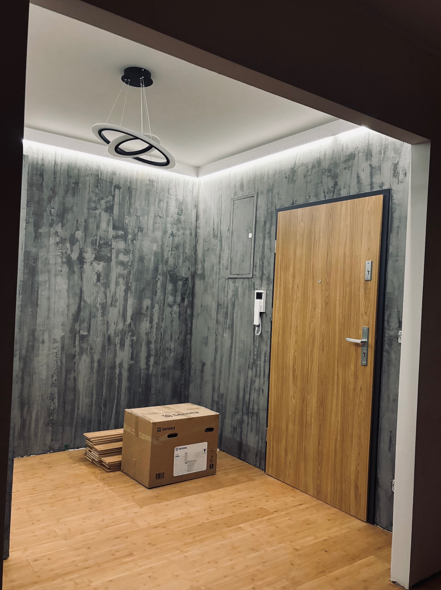 Nowoczesny przedpokój po remoncie: drewniane panele, betonowa ściana, designerska lampa. Widoczna paczka paneli, drzwi wejściowe i oświetlenie LED.