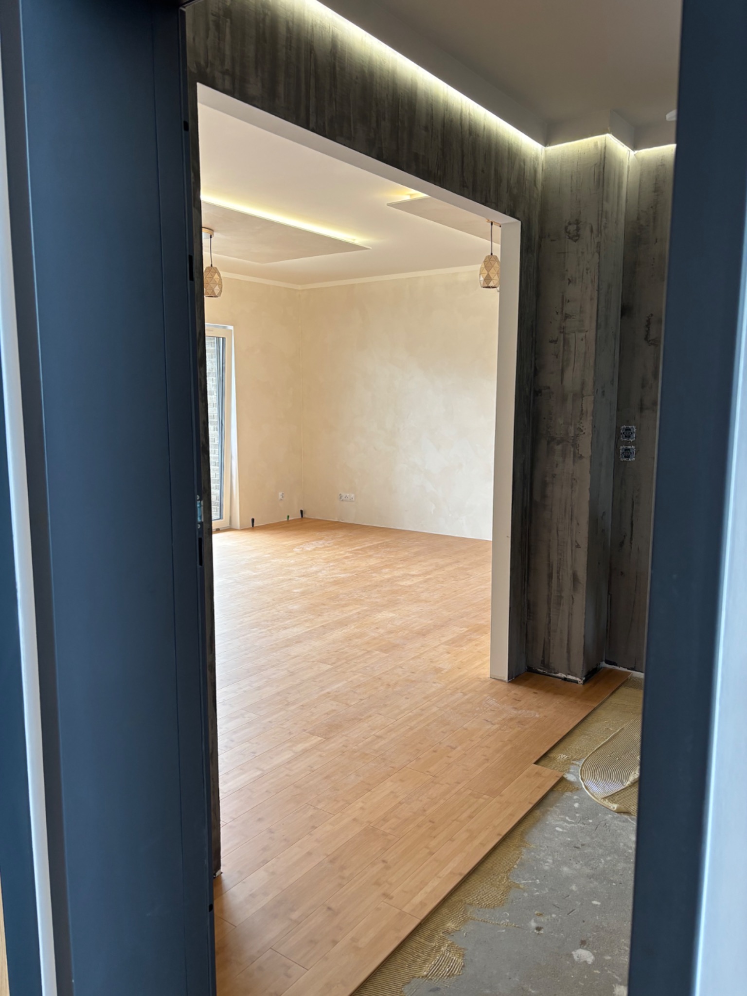 Wnętrze w trakcie układania parkietu, widok zza otwartych drzwi. Część podłogi wykończona, na pozostałej widoczny klej. Ściany z efektem betonu, sufit z oświetleniem.