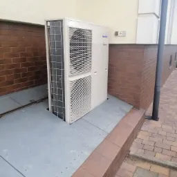 Zewnętrzna jednostka klimatyzacji Panasonic Inverter zamontowana przy budynku z ceglaną podmurówką, na metalowej platformie.