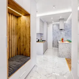 Nowoczesne wnętrze: białe szafy z drewnianą wnęką i szarą poduszką, marmurowe płytki podłogowe, otwarta kuchnia z wyspą i niebieską ścianą akcentującą.