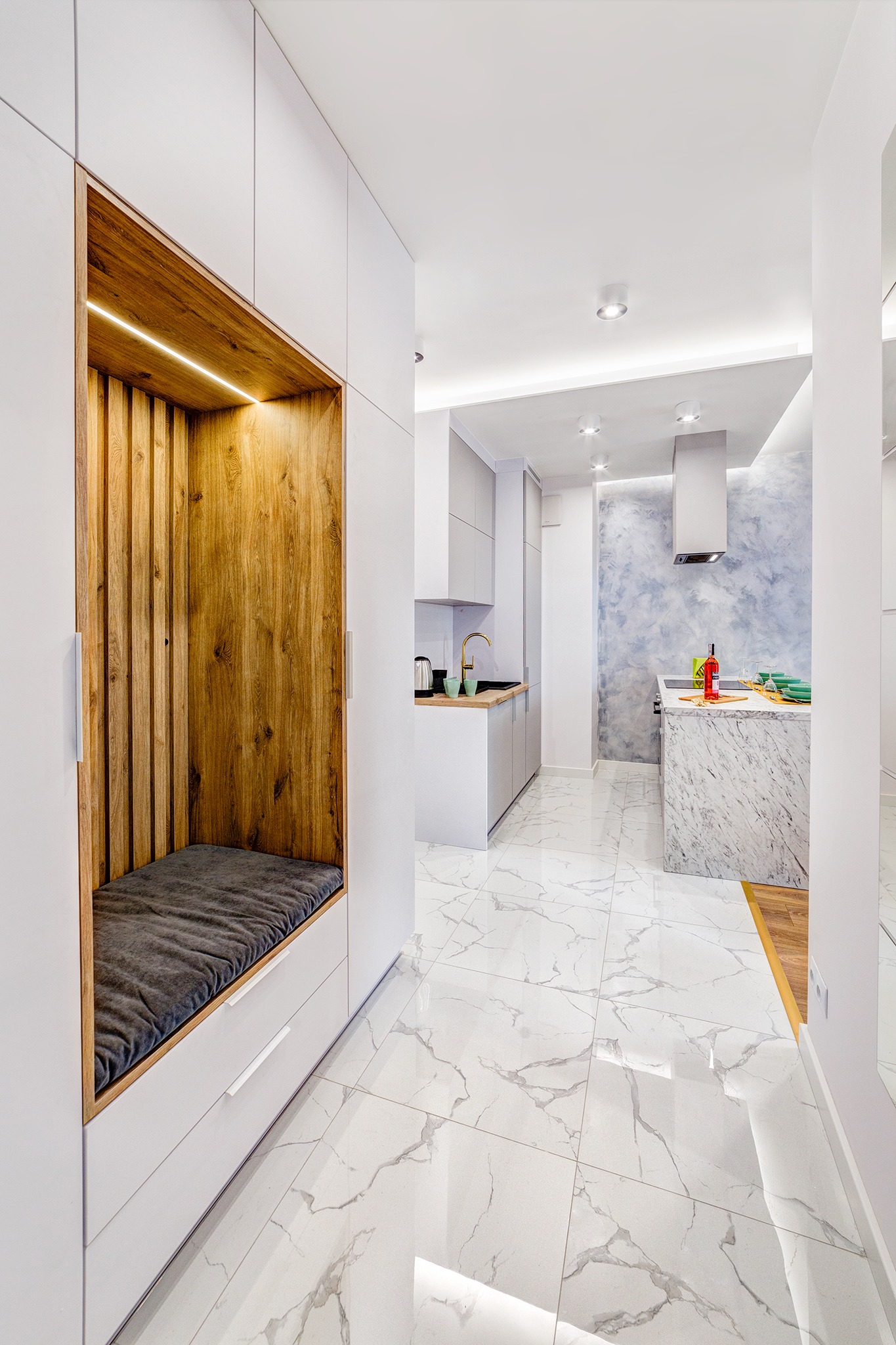 Nowoczesne wnętrze: białe szafy z drewnianą wnęką i szarą poduszką, marmurowe płytki podłogowe, otwarta kuchnia z wyspą i niebieską ścianą akcentującą.