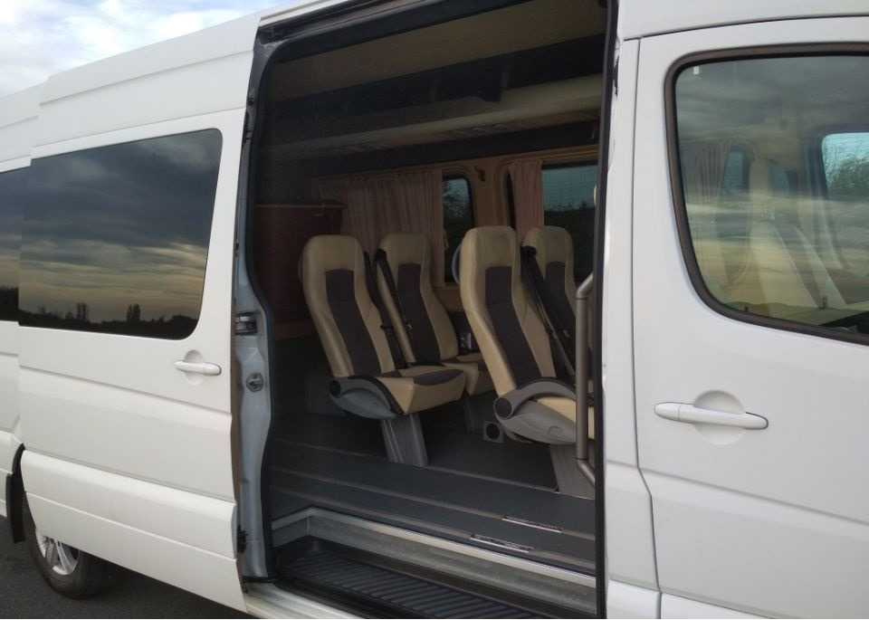 Wnętrze białego busa z beżowo-brązowymi siedzeniami i rozsuwanymi drzwiami, gotowego do przewozu osób.