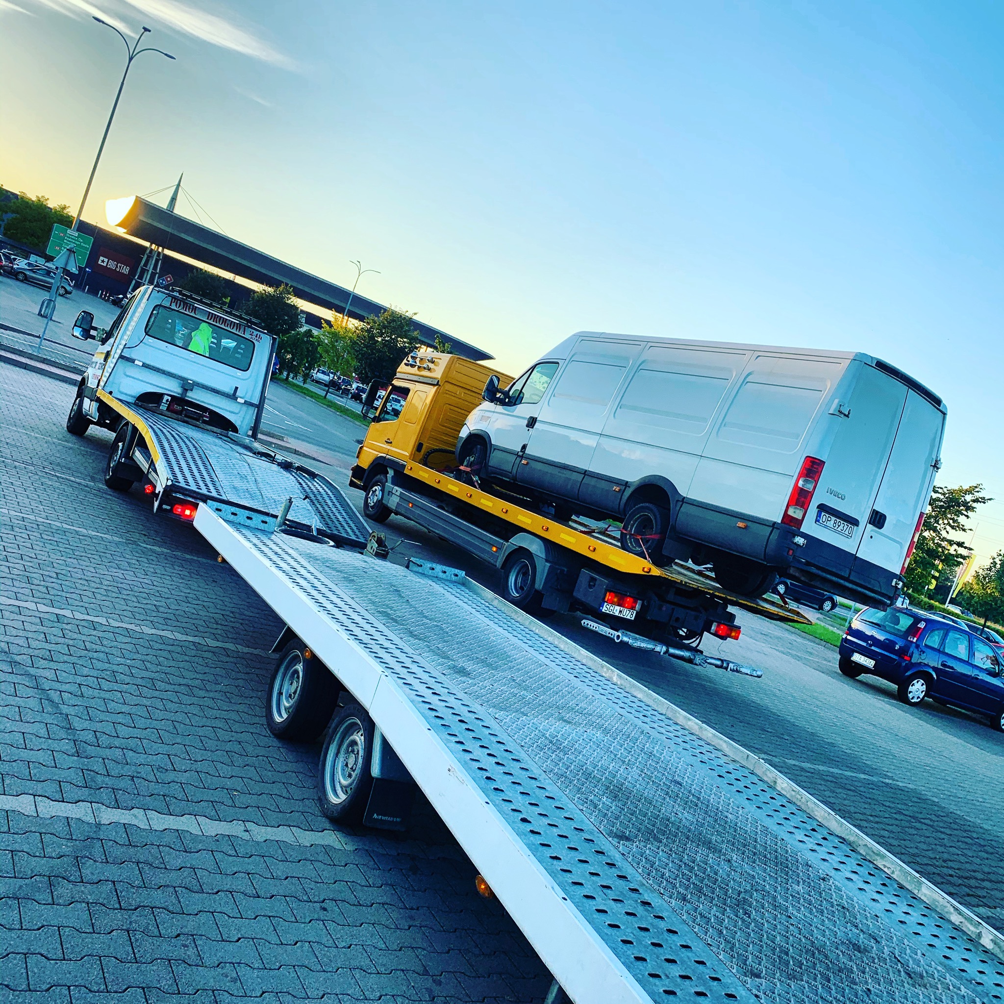 Żółta ciężarówka z platformą transportową przewozi białego dostawczaka na parkingu z kostki brukowej, w tle widoczny budynek z napisem BIG STAR.