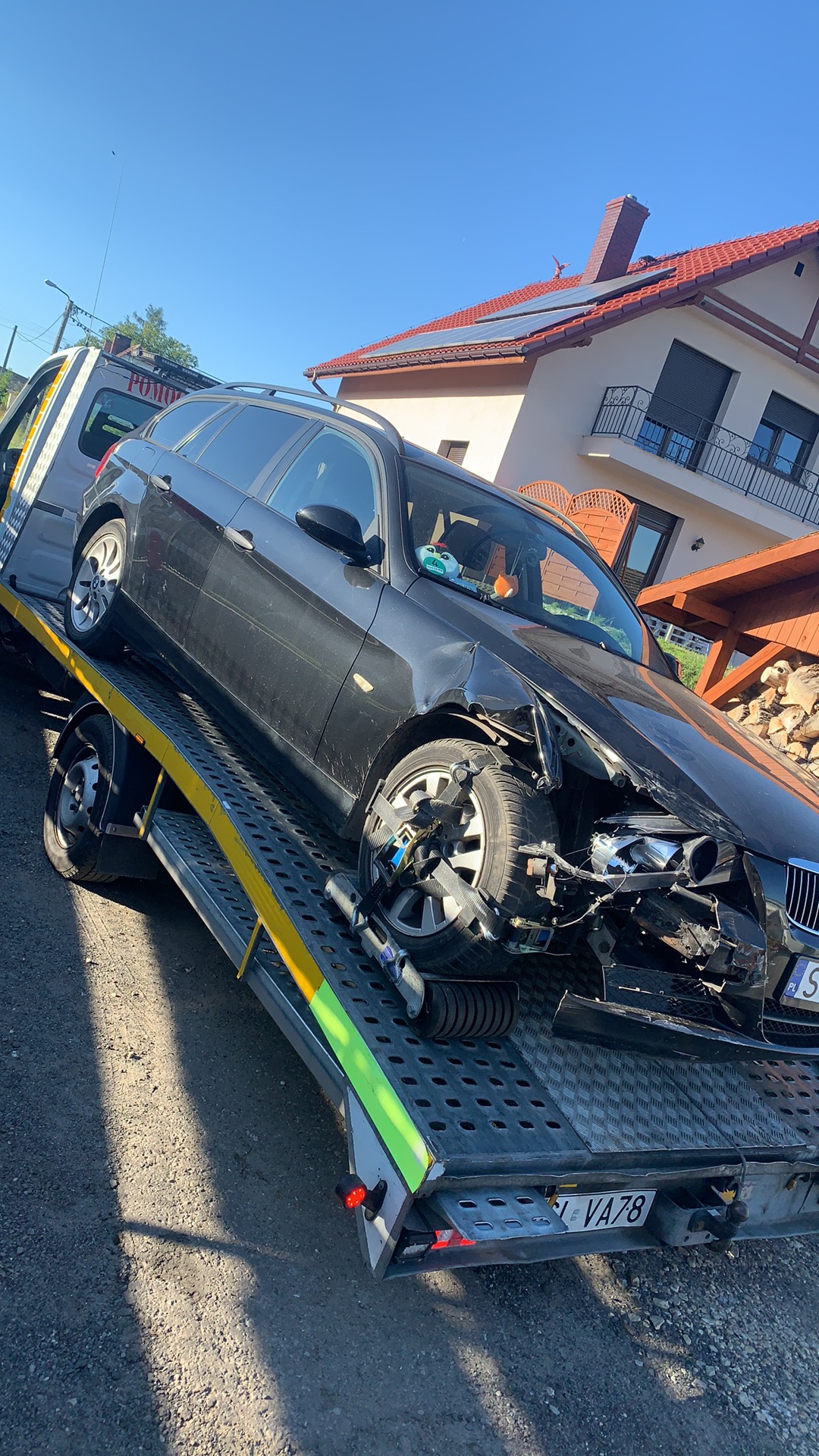 Uszkodzony, czarny samochód osobowy marki BMW na platformie pomocy drogowej, widoczne uszkodzenia przodu pojazdu oraz zabezpieczenia na kołach, w tle dom jednorodzinny z panelami solarnymi na dachu.