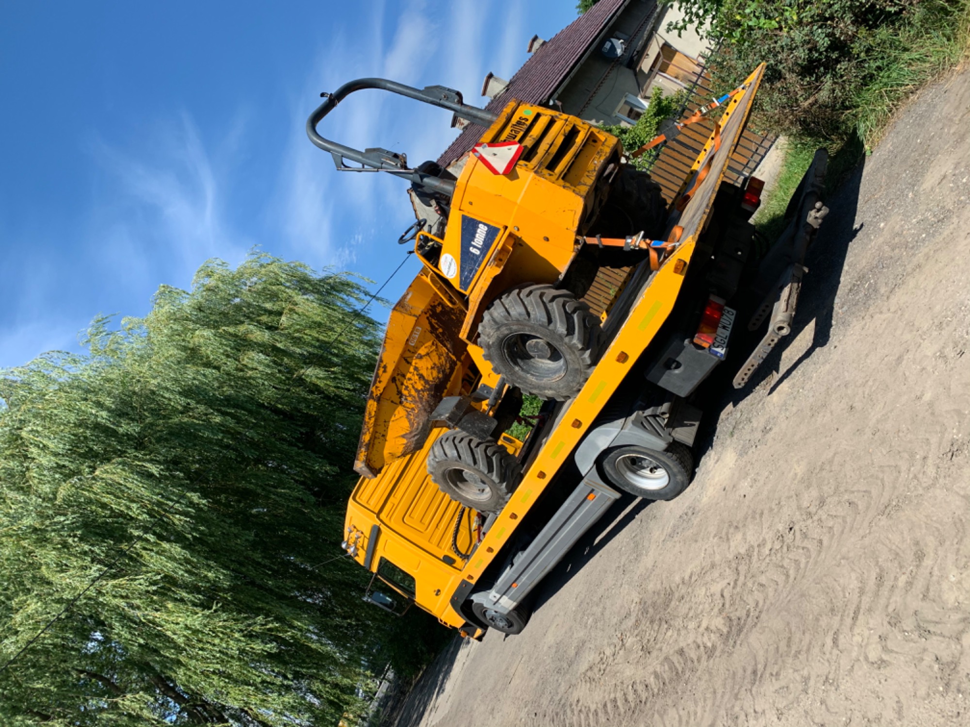 Żółta wywrotka budowlana '6 tonne' zabezpieczona pasami na platformie transportowej, widok z boku na tle błękitnego nieba i zieleni drzew