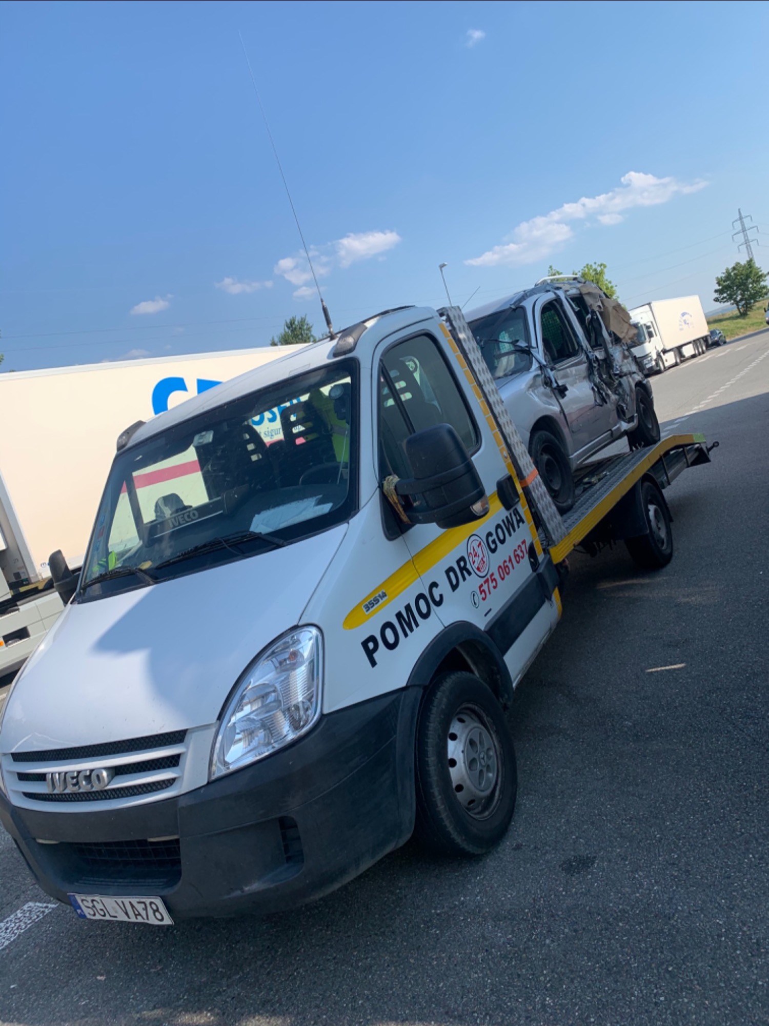 Biała ciężarówka pomocy drogowej Iveco z uszkodzonym srebrnym samochodem osobowym na platformie, widoczne numery kontaktowe 'Pomoc Drogowa' na boku pojazdu, zaparkowana na parkingu w słoneczny dzień.