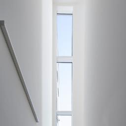 PORT pracownia i studio architektury - Wewnętrzne schody z jasnego kamienia prowadzące do okna pionowego, wpadające światło dzienne, minimalistyczne białe ściany.