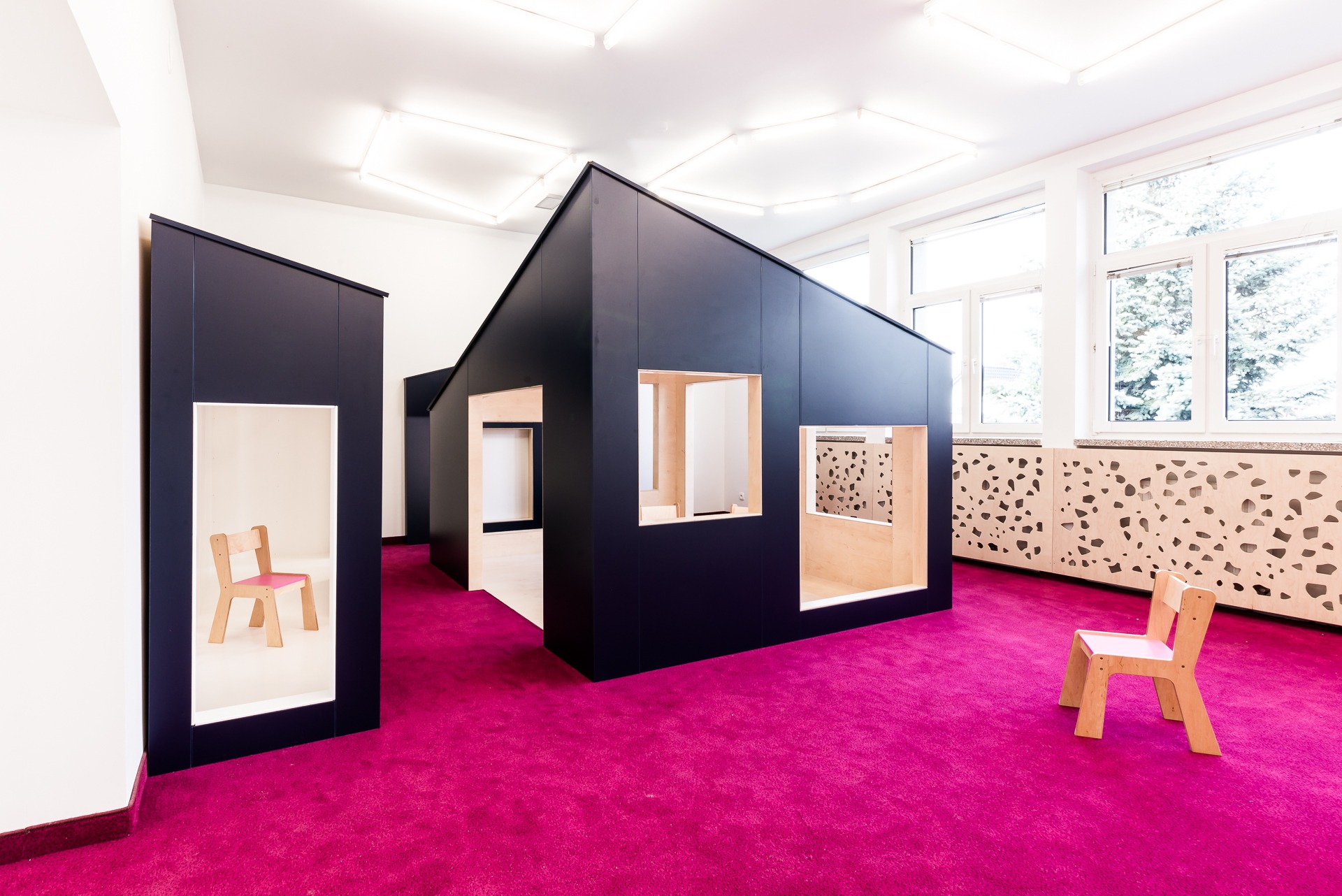 Wnętrze przedszkola: czarne, minimalistyczne konstrukcje w kształcie domków z drewnianymi ramami okiennymi na tle intensywnie różowej wykładziny i ażurowych paneli ściennych.