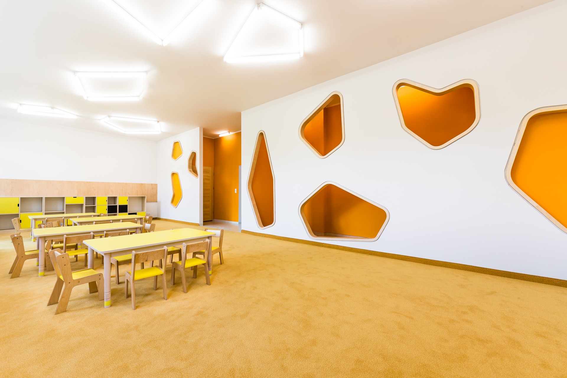 Jasne wnętrze przedszkola z żółtymi meblami i pomarańczowymi wnękami w ścianach, podłoga pokryta wykładziną w odcieniach żółci.