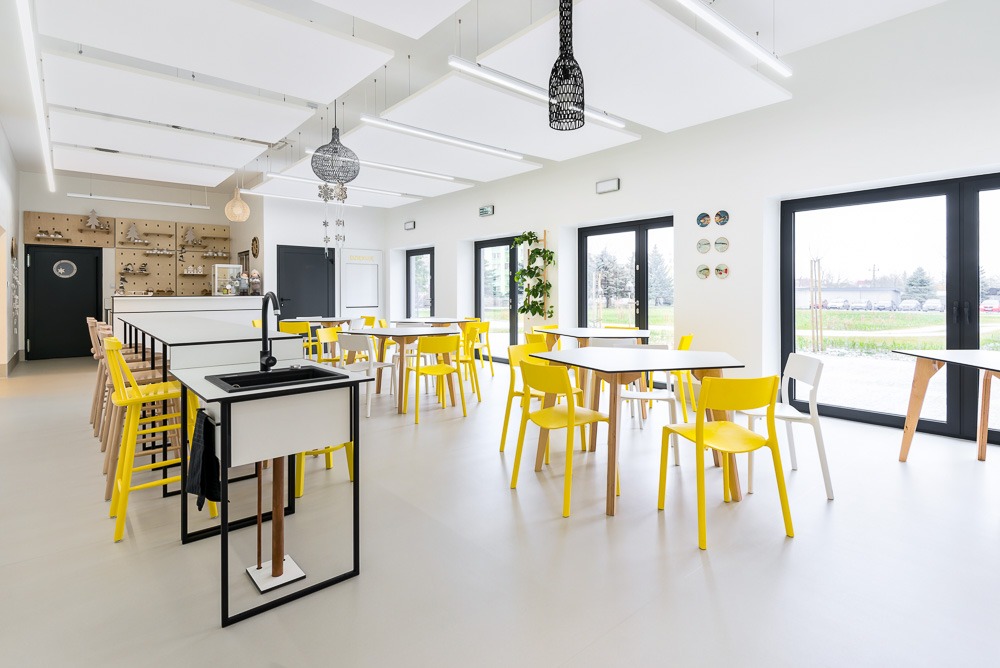 Jasne wnętrze sali szkolnej z białymi ścianami, kwadratowymi stołami i żółtymi krzesłami. Widoczne umywalki z czarnymi kranami oraz dekoracyjne lampy wiszące.