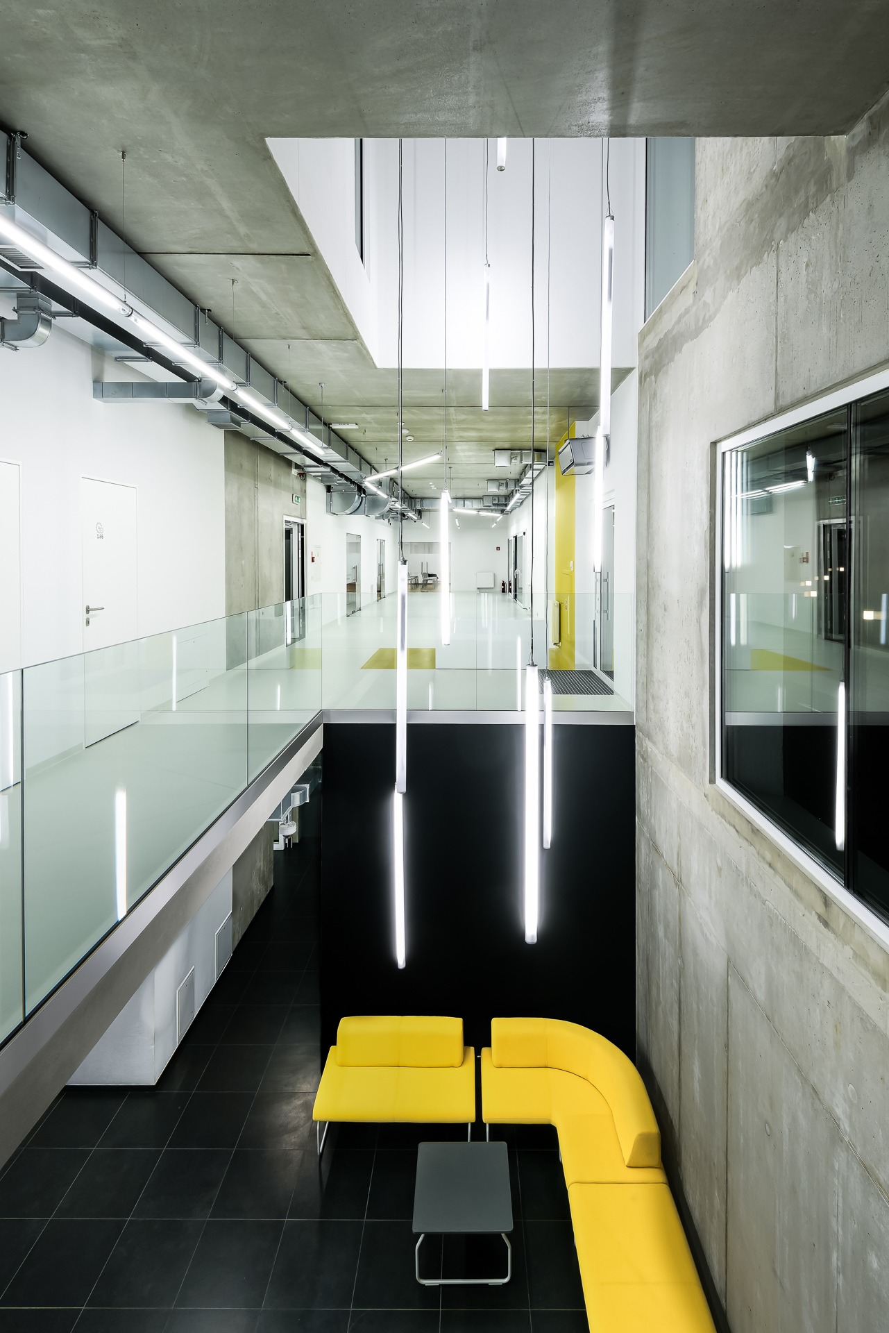 Wnętrze nowoczesnego biurowca z żółtą sofą modułową i szklaną balustradą, widok z góry na hol z podwieszanymi lampami liniowymi.