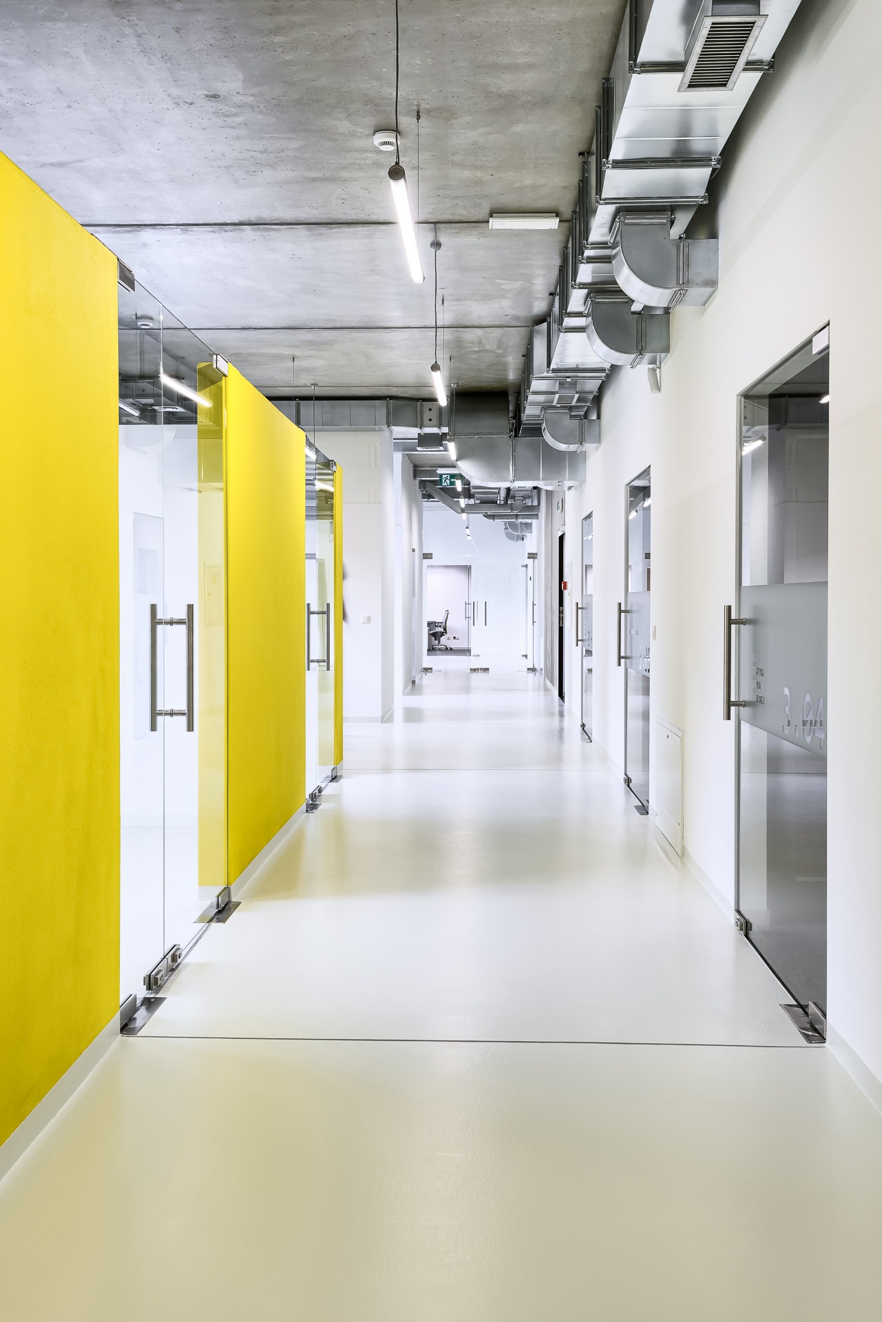 Długi korytarz w nowoczesnym biurowcu z żółtymi ściankami działowymi ze szkła i metalu, widoczne instalacje wentylacyjne pod betonowym sufitem oraz przeszklone drzwi do biur.