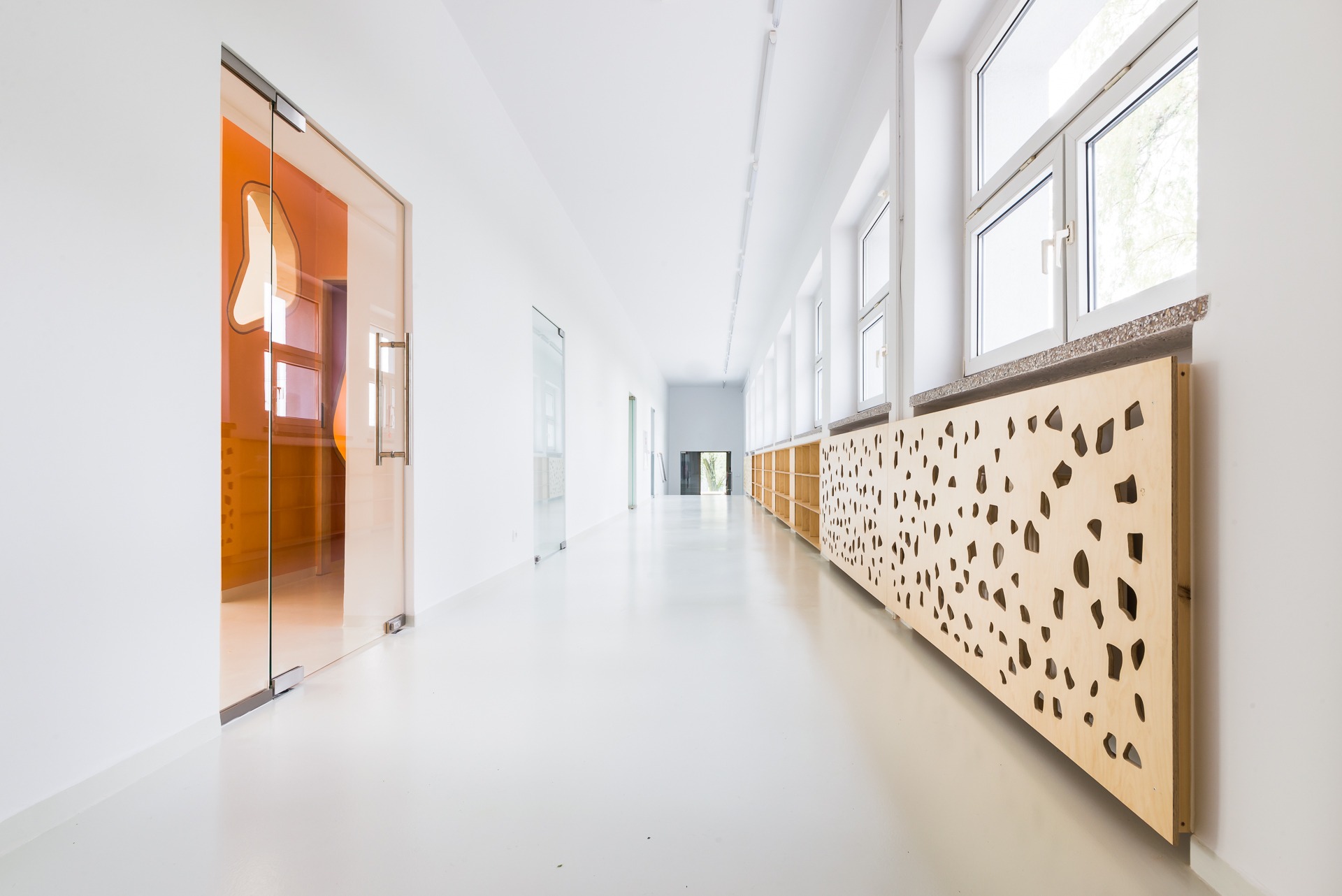 Jasny, przestronny korytarz w przedszkolu z pomarańczowymi akcentami w szklanych drzwiach i ażurowymi osłonami grzejników z jasnego drewna.