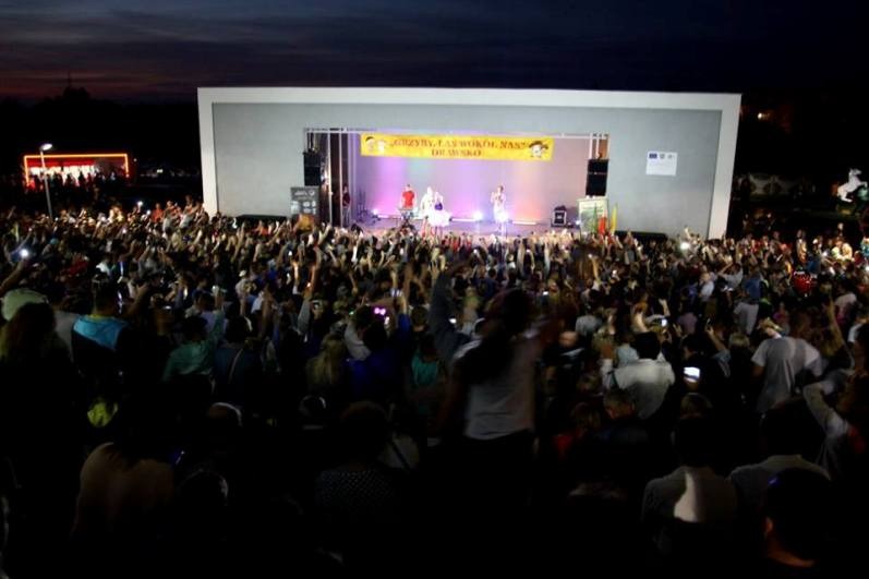 Koncert plenerowy przy zachodzie słońca: zespół na scenie oświetlonej fioletowym światłem, tłum ludzi z podniesionymi rękami, część osób nagrywa teleonami.