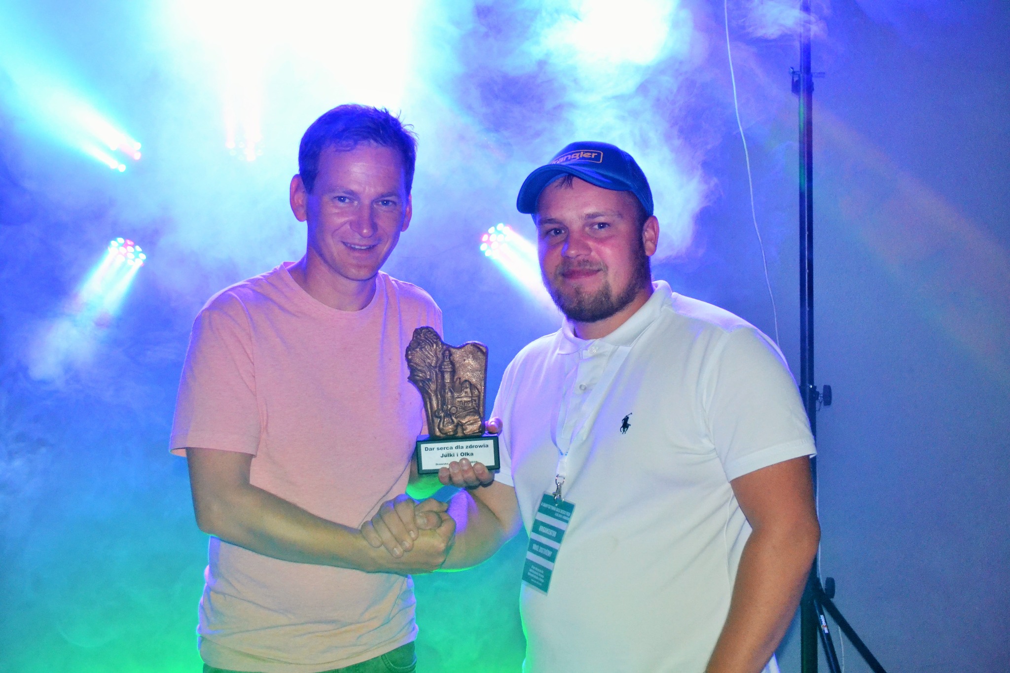 Dwóch mężczyzn w koszulkach wręcza sobie nagrodę na tle niebieskiego oświetlenia scenicznego i dymu.