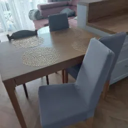 Jasne wnętrze jadalni z drewnianym stołem, szarymi krzesłami z pokrowcami, okrągłymi podkładkami na stole i różową sofą w tle